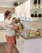 Woman holding a French Bulldog in a kitchen with Feed For Thought Australia packaging on the counter